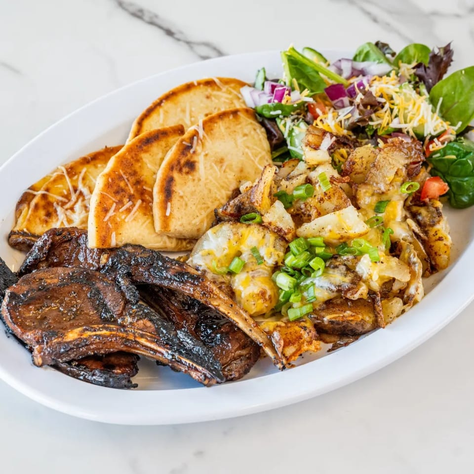 Grilled lamb chops, cheesy potatoes, garlic flatbread, and a fresh side salad on a white plate.